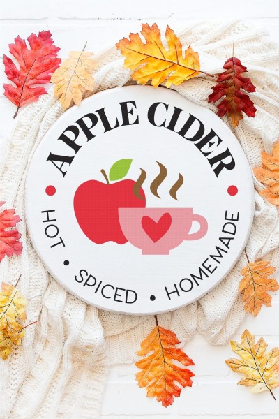 Fall colored leaves sitting next to a round sign that has an apple on it and a coffee mug and says, Apple Cider - Hot-Spiced-Homemade