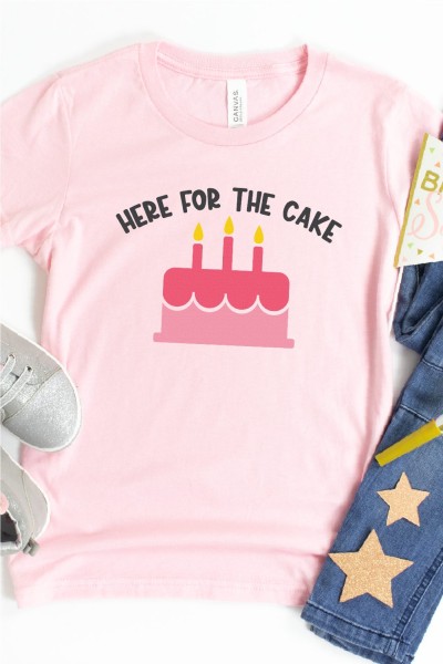 Blue jeans, gray tennis shoes, birthday props and a pink shirt that has a birthday cake on it and the saying Here For The Cake