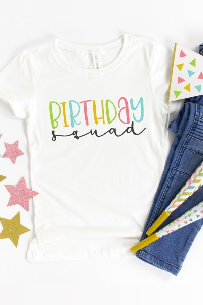 A white shirt decorated with the saying "Birthday Squad" lying next to a pair of blue jeans and birthday decor surrounding it