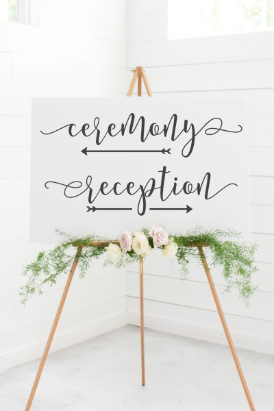 Image of a sign sitting on a tripod decorated with roses and fernlike greenery.  The sign has the text Ceremony with an arrow underneath pointing to the left and the text Reception with and arrow underneath pointing to the right.