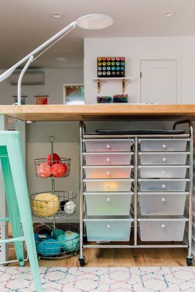 A section of a craft room with a 3 tier wire basket filled with yarn on the floor next to a rolling cart with drawers and a stool.