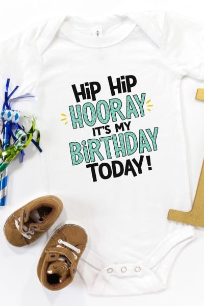 Tiny baby shoes, the number 1, birthday decor and a white onesie that says, Hip Hip Hooray It's My Birthday Today