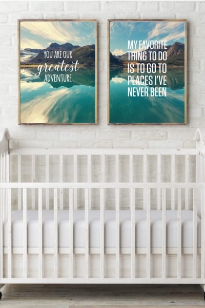 A white baby crib up against a white brick wall with two gold framed pictures hanging above the crib and the pictures are of a mountain and lake and the sayings, "You Are Our Greatest Adventure" and "My Favorite Thing to do is to go to Places I've Never Been"