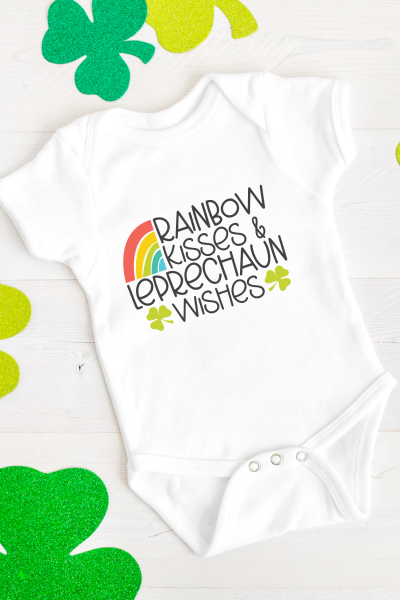 White baby onesie with a rainbow and three leaf clovers on it and the saying Rainbow Kisses and Leprechaun Wishes